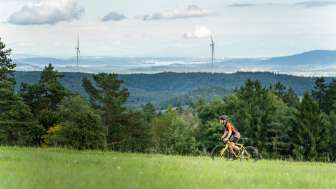 A man is riding his yellow gravel bike along a dirt road. In the background, there is a sweeping view of forests, wind turbines, and a lake.