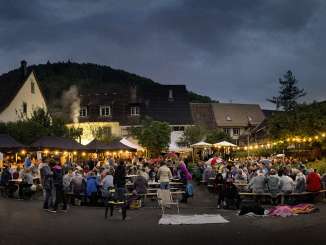 Many people are sitting at wooden tables in a square. A string of lights creates a cozy atmosphere.