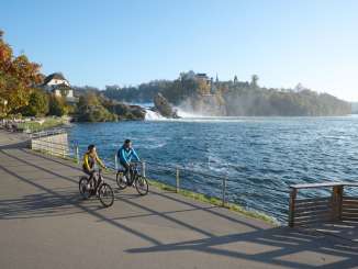 RheinWelten Two cyclists on the banks of a river. A waterfall can be seen in the background. On the other bank is a castle.