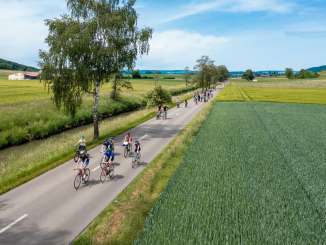 Many cyclists are riding on a road that is closed to cars. The road runs alongside a stream and fields.