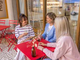 Three friends are sitting at a small red garden table in an arbor in the old town. They all have a glass of wine in their hands and are chatting.