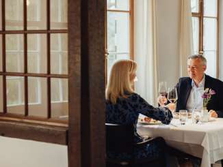 A couple are sitting at a table set in white. They are enjoying a gourmet meal and toasting with a glass of red wine.