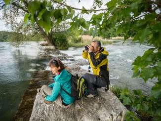 Two hikers are using binoculars to watch animals by the water in Schaffhausen Nature Park.