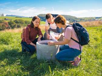 Savurando - culinary treasure hunt Three people kneel around a concrete pipe in a meadow. They are solving a riddle.