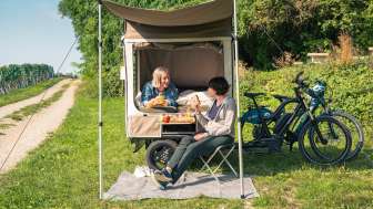 In a meadow next to vineyards, there is a cargo bike with an unfolded tent. A person is lying in bed and sitting on a camping chair in front of it.