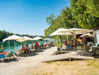 Entlang des Rheins stehen mehrere bunte Tische und Stühle mit Sonnenschirmen. Am Weg steht ein Wagen als Pop-Up Restaurant.