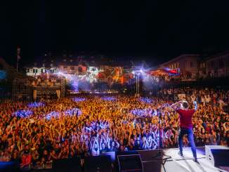 View from the stage with the singer onto a square in the middle of the old town filled with people. The facades of the houses are illuminated with a light show.