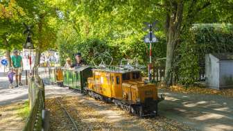 A small miniature train with an orange locomotive drives through a park with a train driver and a child as a passenger.