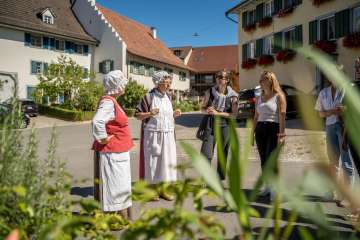 Two costumed ladies are telling stories. The other people are listening and laughing. Old houses stand in the background.