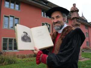 A man in a black cloak holds up a Buch . An old painted portrait can be seen on the left-hand page of the book. A red statue and a red house façade can be seen in the background.