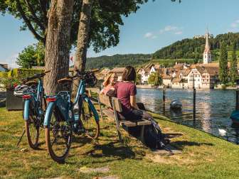 Two friends are sitting on a wooden bench by the Rhine, with a view of Stein am Rhein. Two e-bikes are parked behind them.