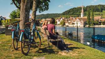 Two friends are sitting on a wooden bench by the Rhine, with a view of Stein am Rhein. Two e-bikes are parked behind them.