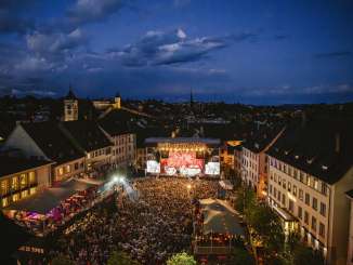A square between houses in the old town. The square is filled with people. In front is a stage with the inscription "Stars in Town".