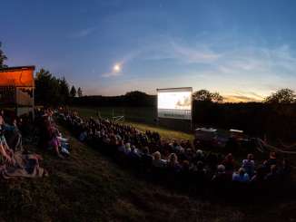 A large group of people are sitting on a meadow on a gentle slope, watching a movie on a big screen.