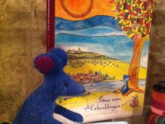 A felted blue Mai stands in front of a boo with the title "Titus von Hohenklingen".