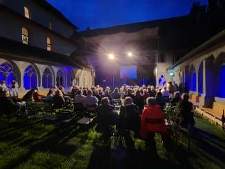 There is seating in an inner courtyard on the lawn. A temporary stage is set up. Around 50 people are in the audience.