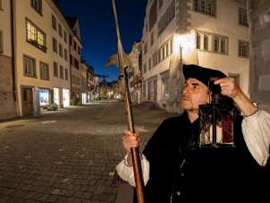 It is dusk in Stein am Rhein. A person dressed as a night watchman stands in the middle of the old town of Stein am Rhein. He holds a lantern in one hand and a halberd in the other.