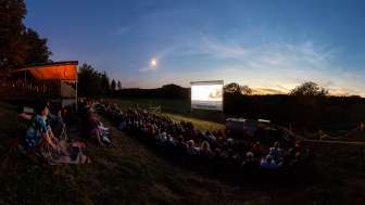 A large group of people are sitting on a meadow on a gentle slope, watching a movie on a big screen.