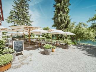 Terrace Gasthof Schupfen Wooden chairs and tables are set and white parasols provide shade. The restaurant terrace is located directly on the Rhine, surrounded by greenery.