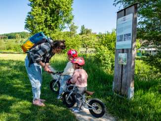 Biberweg Thayngen An adult woman with two children on a path next to a river. Both children are sitting on a balance bike. Next to them is a plaque with a picture of a beaver.
