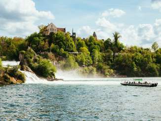 A green boat sails towards the waterfall in the Rhine Falls basin. There are many people on board.