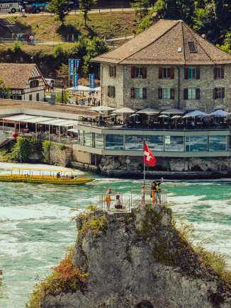 Middle rock Rhine Falls Several people and a Swiss flag stand on the rock in the middle of the waterfall. Behind the rock you can see the Schlössli Wörth.