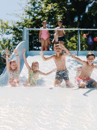 Four children slide down a wide slide together in an outdoor pool.