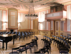 Classic Rüden A grand piano stands in a hall with a stucco ceiling. Chairs in concert chairs stand around the grand piano.