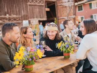 A group of friends is sitting at a banquet table decorated with flowers. A barn can be seen in the background. The friends are toasting with a glass of wine.