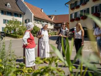 Two costumed ladies are telling stories. The other people are listening and laughing. Old houses stand in the background.