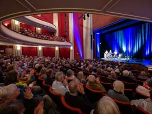 Erzählzeit without borders A hall filled with people clapping their hands. An illuminated stage with three people on it, a sofa and an armchair.