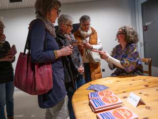 An author is sitting at a table with her books. Four people are standing in line in front of her. The author hands one person her Buch.