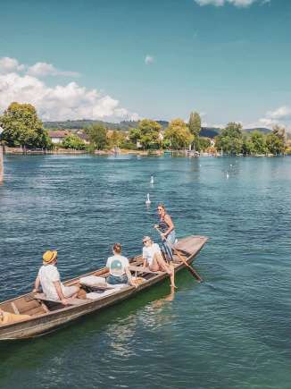 Three people and a dog are sitting in the Weidling. The wooden boat drifts leisurely down the Rhine. A woman is standing, holding an oar and steering the boat.