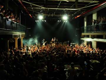 A hall filled with people. There is a balcony to the left and right. In front is a stage with a band playing a concert.