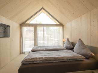 A double bed with gray bed linen in a wooden hut. The large window has a translucent curtain.