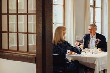 A couple are sitting at a table set in white. They are enjoying a gourmet meal and toasting with a glass of red wine.