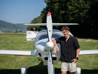 Lukas Britschgi, European figure skating champion, leans against a glider. He is wearing a black short-sleeved shirt and beige shorts.
