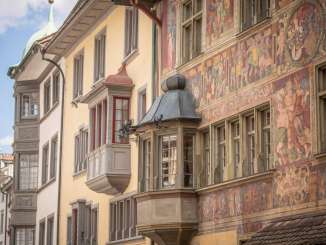 A view of several house facades featuring oriel windows. One facade is light yellow, while another is richly adorned with paintings.