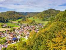 View from the forest of a village surrounded by autumnal, forest-covered hills. There is a church just outside the village.