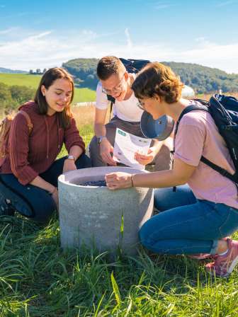 Three people kneel around a concrete pipe in a meadow. They are solving a riddle.