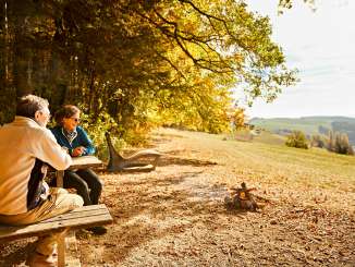 A couple is sitting on a wooden bench next to a fire pit. The forest is ablaze with autumn colors.