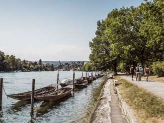 Four hikers walk along the banks of the Rhine. There are various weidlings in the Rhine. The Munot fortress and the town of Schaffhausen can be seen in the distance.