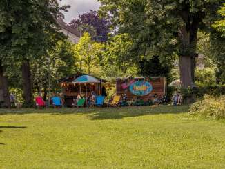 In a park, there is a small wooden house and a trailer that serve as a pop-up café. In front of it are lots of colorful deckchairs and wooden tables with chairs.