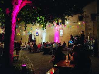 Festival benches in an illuminated courtyard. The benches are filled with people. A kleine Bühne has been set up. A musician is playing on it.