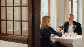 A couple sits at a white-clothed table. They have a gourmet menu in front of them and toast with a glass of red wine.
