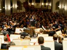 An orchestra on a church stage. The conductor sets the beat and the pews are filled with concertgoers.