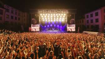 Stars in the city Many people with their arms in the air are at a concert with a band on a large stage.
