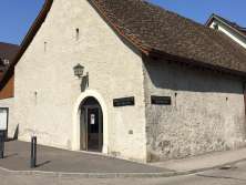 An older house with a white stone façade and brown roof. There are no windows to be seen, just small peepholes. The sign at the entrance readsTrottentheater Neuhausen".