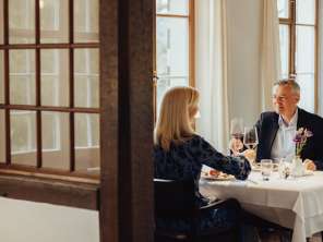 A couple are sitting at a table set in white. They are enjoying a gourmet meal and toasting with a glass of red wine.