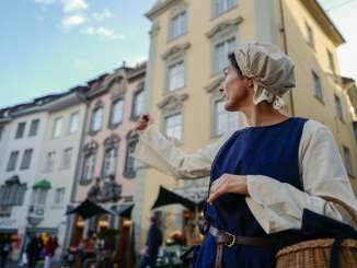 The person dressed as a peddler points to a landmark with her arm.